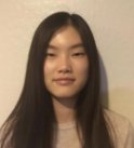 A young asian woman with long dark brown hair wears a beige t-shirt and smiles in front of a white wall.